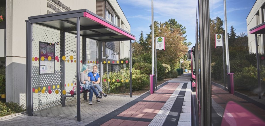 zwei Gasbusse der koveb halten an einer Haltestelle an zwei hintereinander stehende Gasgelenkbusse der koveb halten an einer Haltestelle der koveb an und zwei Fahrgäste warten währenddessen in dem Wartehäuschen der Bushaltestelle.