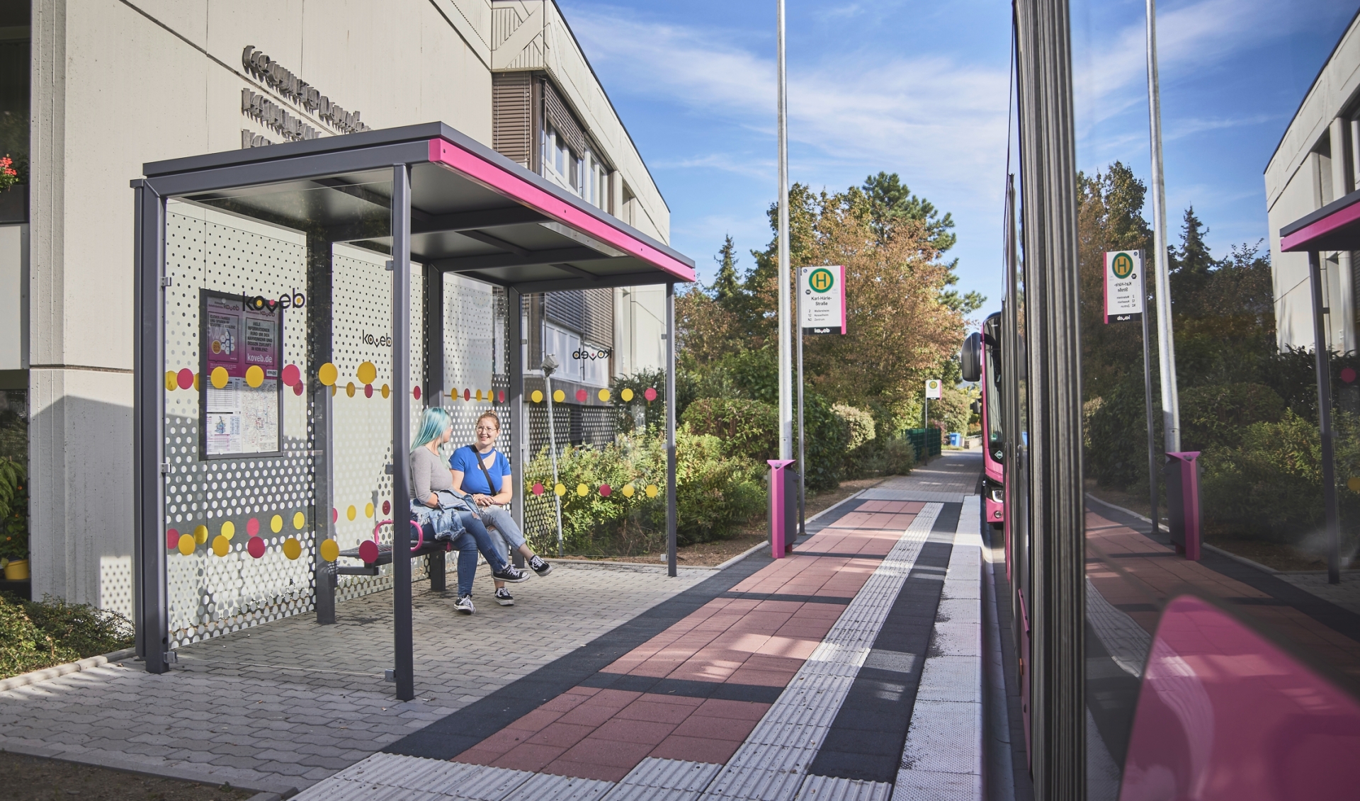 zwei hintereinander stehende Gasgelenkbusse der koveb halten an einer Haltestelle der koveb an und zwei Fahrgäste warten währenddessen in dem Wartehäuschen der Bushaltestelle.