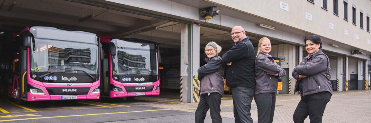 Drei Busfahrerinnen und ein Busfahrer stehen in lockerer Pose vor einer Bushalle der koveb