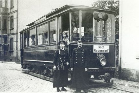 historischer OBus auf Schienen mit davor stehendem Schaffner und Fahrkartenkontrolleur in langen Mänteln und Schirmmütze