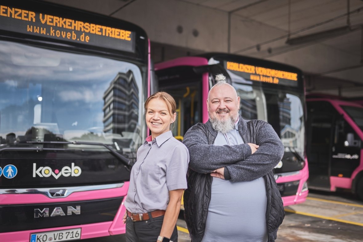 Eine Busfahrerin und ein Busfahrer stehen vor koveb-Bussen und lächeln.