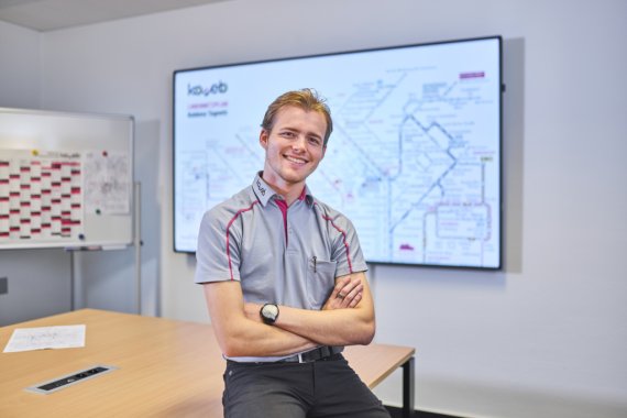 Ein Auszubildender der koveb lehnt an einem Tisch - im Hintergrund ist ein Bildschirm mit dem Liniennetz der koveb zu sehen.