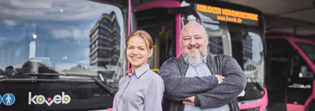 Eine Busfahrerin und ein Busfahrer stehen Schulter an Schulter vor einigen Bussen der koveb und lächeln