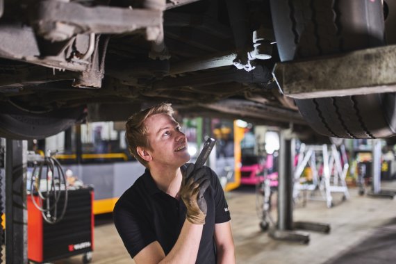 Arbeiten als Mechatroniker bei der koveb Ein Werkstatt-Mitarbeiter der koveb untersucht den Unterboden eines hochgebockten koveb-Busses mit einer Taschenlampe.