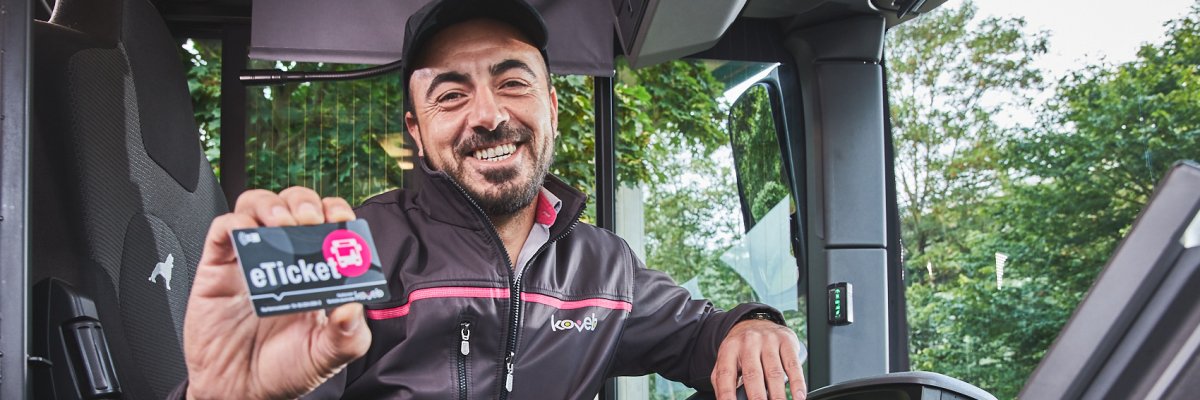 Ein freundlicher Busfahrer der koveb lächelt und hält ein eTicket in die Kamera