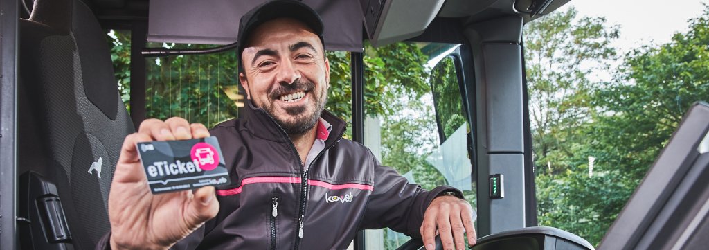 Ein freundlicher Busfahrer der koveb lächelt und hält ein eTicket in die Kamera